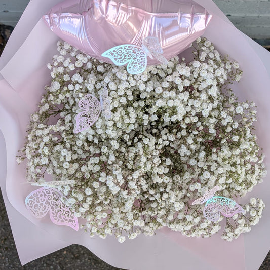 A bouquet of white baby's breath flowers wrapped in pale pink paper, with a metallic pink balloon and small decorative butterfly embellishments, photographed outdoors against a painted brick wall.