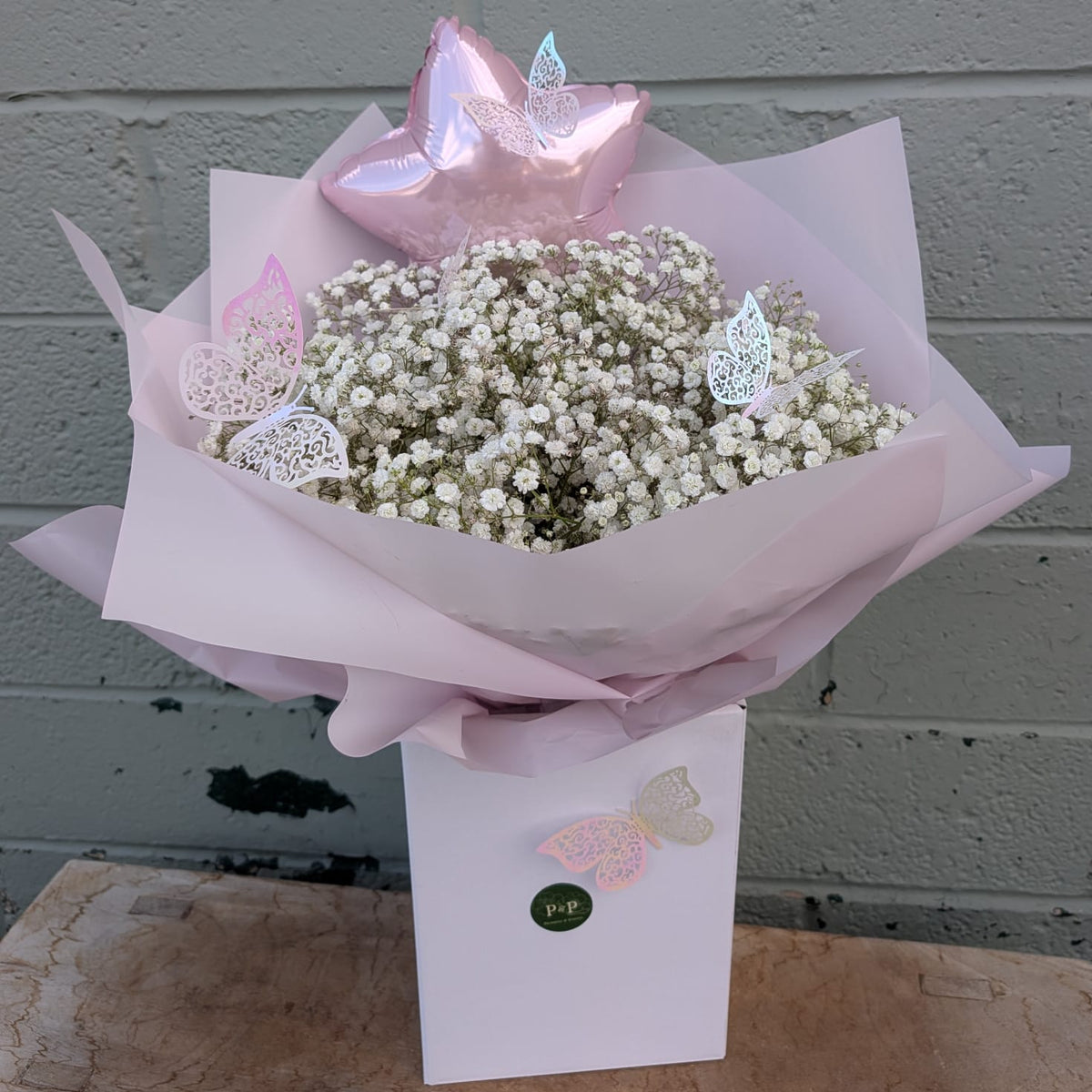 A baby's breath flower bouquet wrapped in pale pink tissue paper, topped with a metallic pink star balloon and butterfly decorations, presented in a white box against a gray brick wall.
