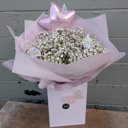 A baby's breath flower bouquet wrapped in pale pink tissue paper, topped with a metallic pink star balloon and butterfly decorations, presented in a white box against a gray brick wall.