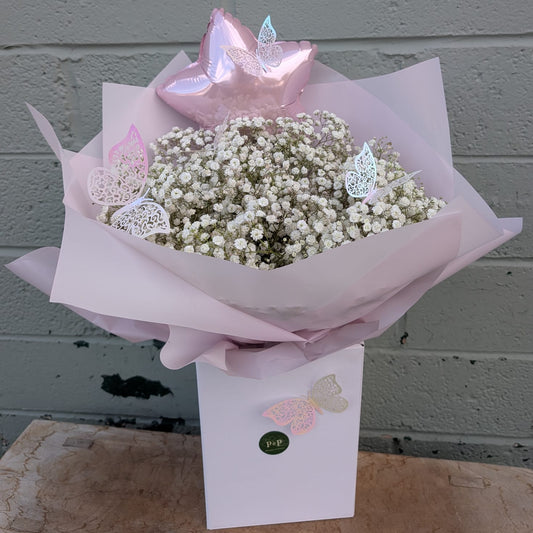 A baby's breath flower bouquet wrapped in pale pink tissue paper, topped with a metallic pink star balloon and butterfly decorations, presented in a white box against a gray brick wall.