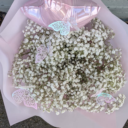 A bouquet of white baby's breath flowers wrapped in pale pink paper, with a metallic pink balloon and small decorative butterfly embellishments, photographed outdoors against a painted brick wall.