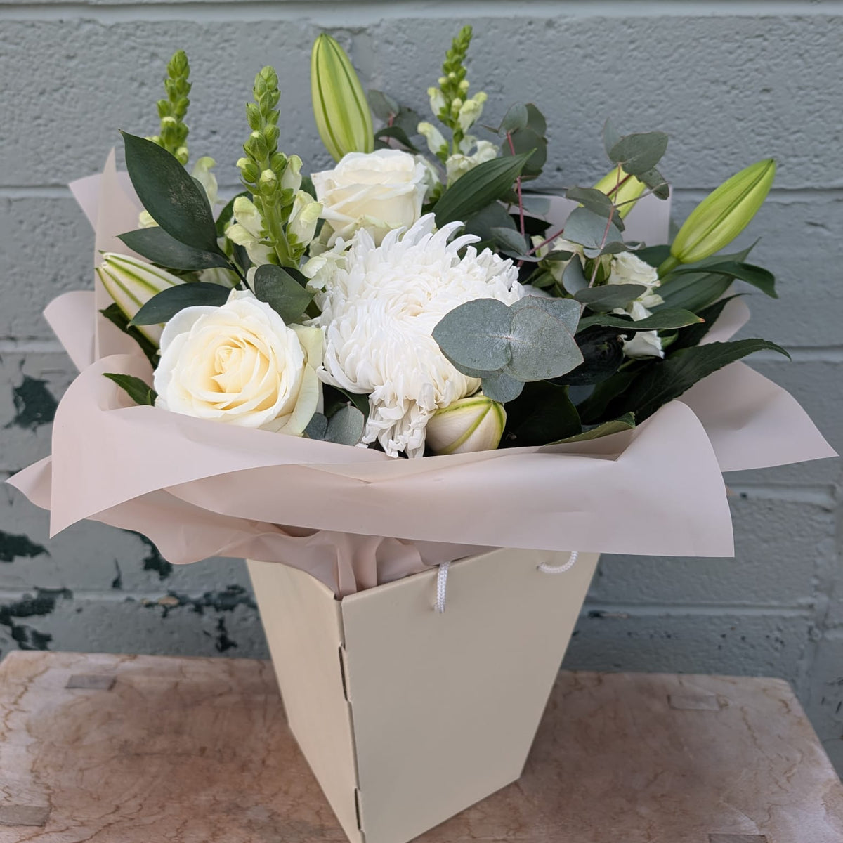 A floral bouquet with white roses, white chrysanthemums, green lily buds, and green foliage, wrapped in pale blush paper and placed in a cream-colored box against a gray painted brick wall.