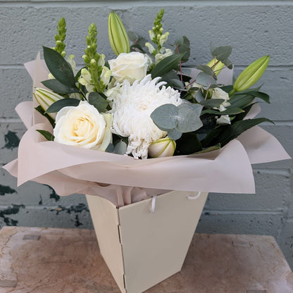 A floral bouquet with white roses, white chrysanthemums, green lily buds, and green foliage, wrapped in pale blush paper and placed in a cream-colored box against a gray painted brick wall.