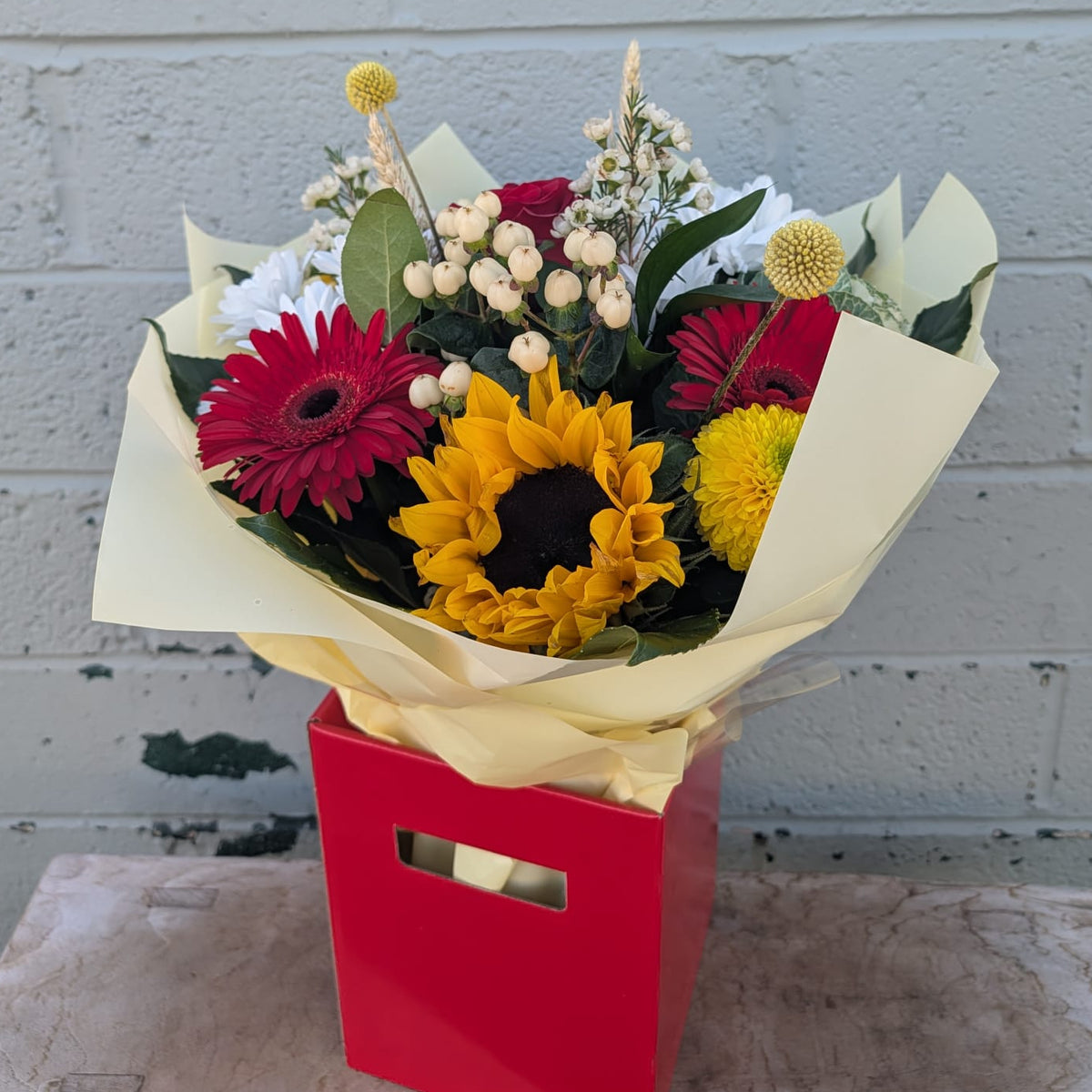 A vibrant mixed flower bouquet featuring yellow sunflowers, red gerbera daisies, red and yellow chrysanthemums, and green foliage, wrapped in cream paper and presented in a red box against a white painted brick wall.