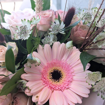 A close-up of a floral arrangement featuring a large light pink gerbera daisy with a dark center, surrounded by blush pink roses, green foliage, and soft white flowers.