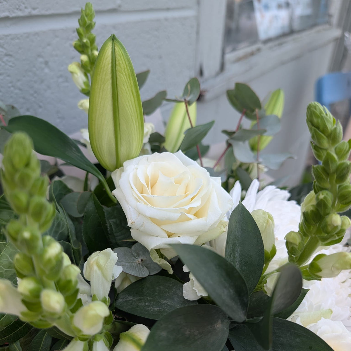 A close-up of a floral arrangement featuring white roses, pale green lily buds, white snapdragons, and dark green eucalyptus leaves, set against a light gray brick wall background.