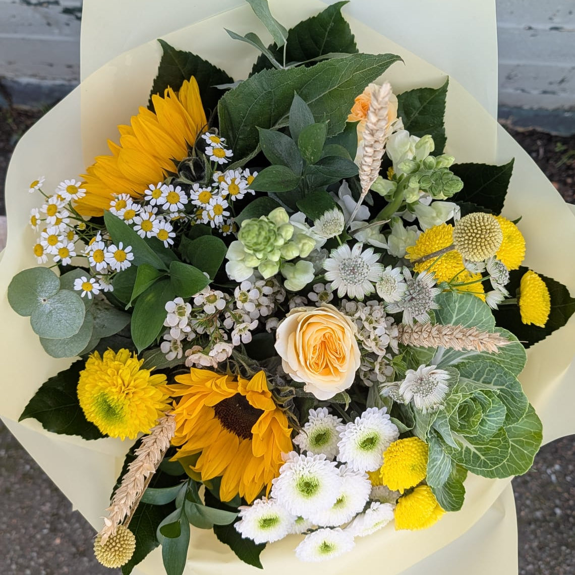 A mixed flower bouquet wrapped in pale yellow paper, featuring sunflowers, white daisies, a peach rose, green foliage, and yellow pom-pom chrysanthemums, photographed outdoors against a white brick wall.