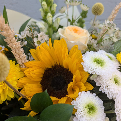 A close-up of a floral arrangement featuring a large yellow sunflower, white daisy-like flowers, green foliage, and a pale peach rose, photographed against a light gray background.